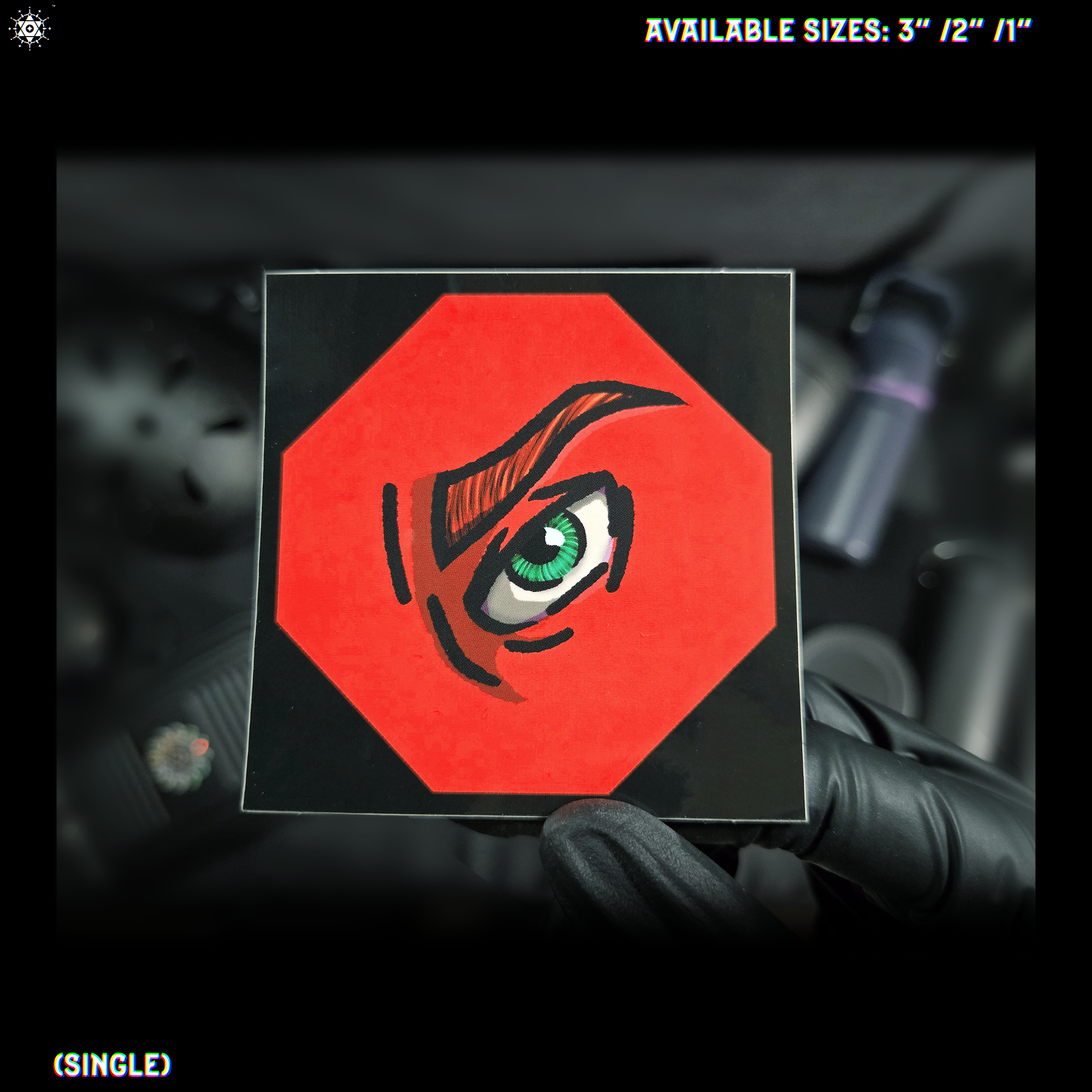 Video of hand holding square shaped sticker design depicting an eye; green iris, red/orange skin and eyebrows, black line art and borders shaped as a octagon.